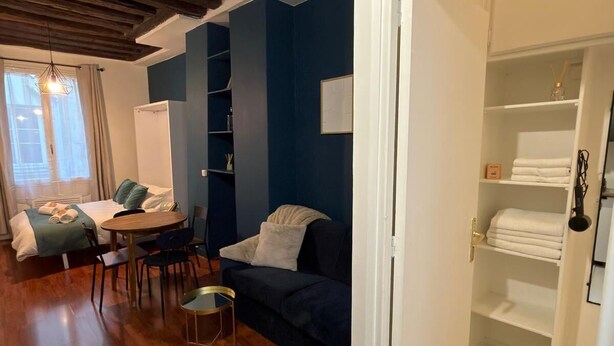 Et værelse med træloft, blå væg, seng, sofa, spisebord med stole og et hvidt skab med hylder.