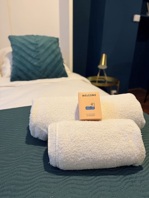 Ein ordentlich bezogenes Bett mit einem 'WELCOME'-Schild und einem Handtuch auf einer gemusterten, grünen Bettdecke.