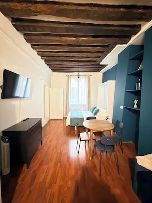 Ein Zimmer mit Holzboden, einem Bett mit blauer Kopfteil, einem an der Wand montierten Fernseher und einem Schreibtisch mit Stuhl.