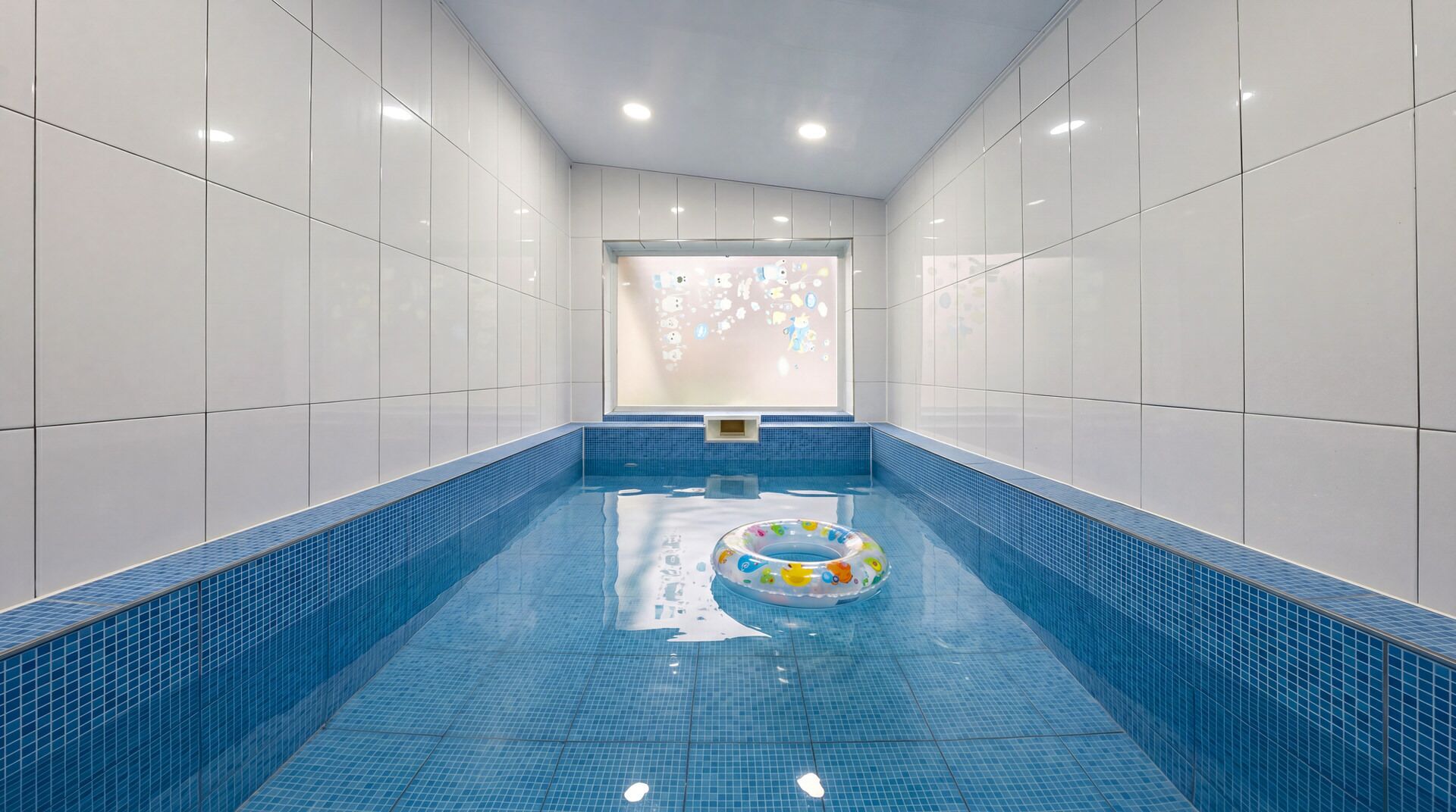 A swimming pool with a blue tiled floor and walls, a floating ring, and a window with a view of the sky.