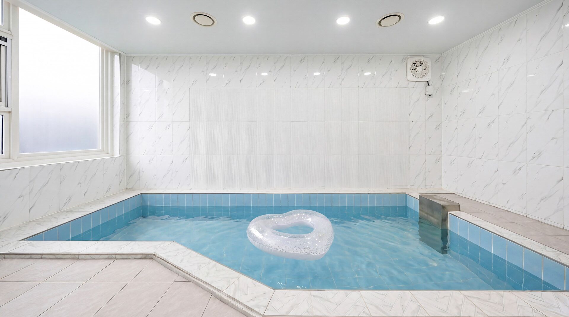 A rectangular indoor swimming pool with a heart-shaped inflatable ring, surrounded by white marble tiles and a window on one side.
