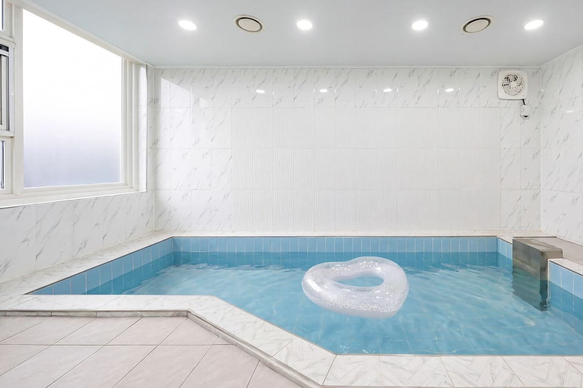 Una piscina interior rectangular con un flotador inflable con forma de corazón, rodeada de baldosas de mármol blanco y una ventana en un lateral.