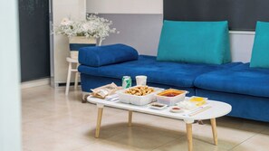 A dining table with food containers, a can, and a cup in a modern living room with a blue sofa and a flower vase.