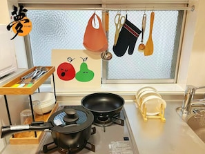 A kitchen with a gas stove, a frying pan, a pot, a wooden tray with utensils, and a window with hanging kitchen items. - YUME Yume Matsuyacho 301 ｜Namba/Shinsaibashi｜Walking distance from Matsuyacho station｜Family welcome (大阪市)
