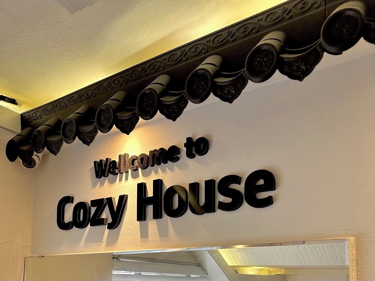 Ein Schild mit dem Text 'Welcome to Cozy House' über einer Zierdecke.