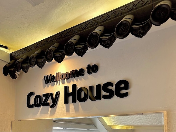 Placa com o texto 'Welcome to Cozy House' acima de um teto decorativo.