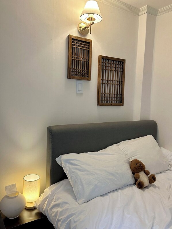 A neatly made bed with white bedding, a teddy bear on the pillow, a bedside lamp, and two wall-mounted wooden panels.
