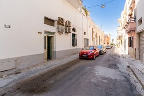 Une rue étroite, bordée de bâtiments des deux côtés, une voiture rouge garée à droite et un ciel bleu dégagé.