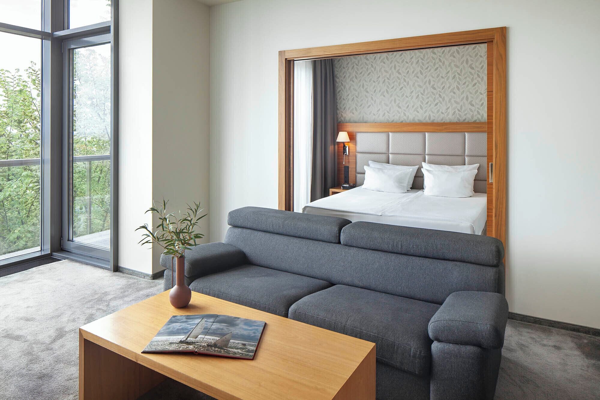 Une chambre d’hôtel avec un lit, un canapé gris, une table basse en bois et une fenêtre donnant sur des arbres.