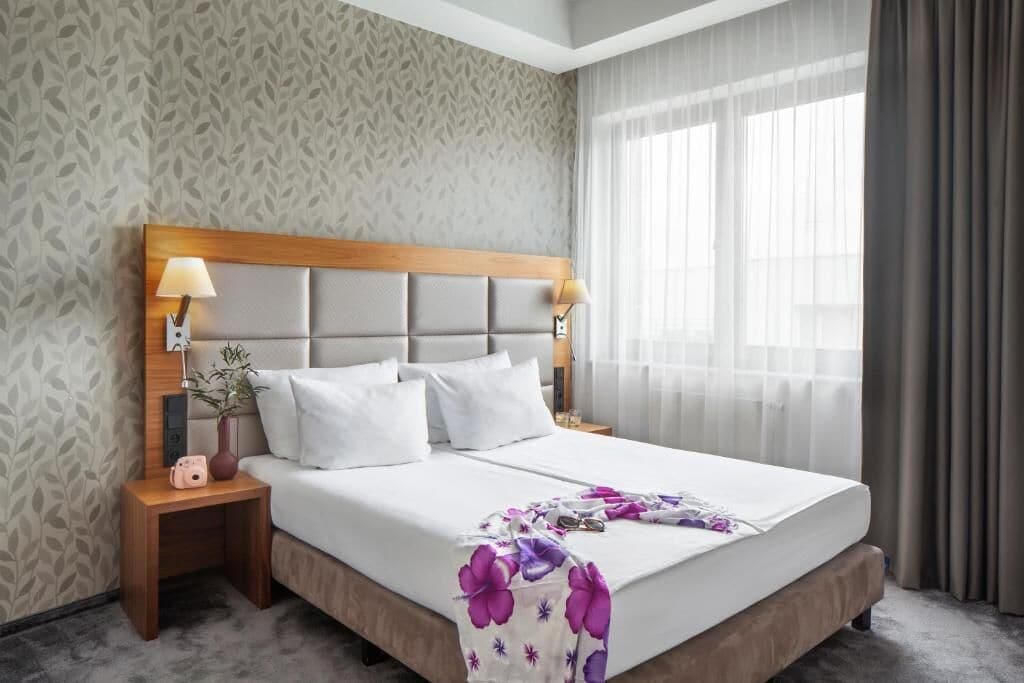 A hotel room with a bed, nightstands, lamps, a window with curtains, and a patterned wall.