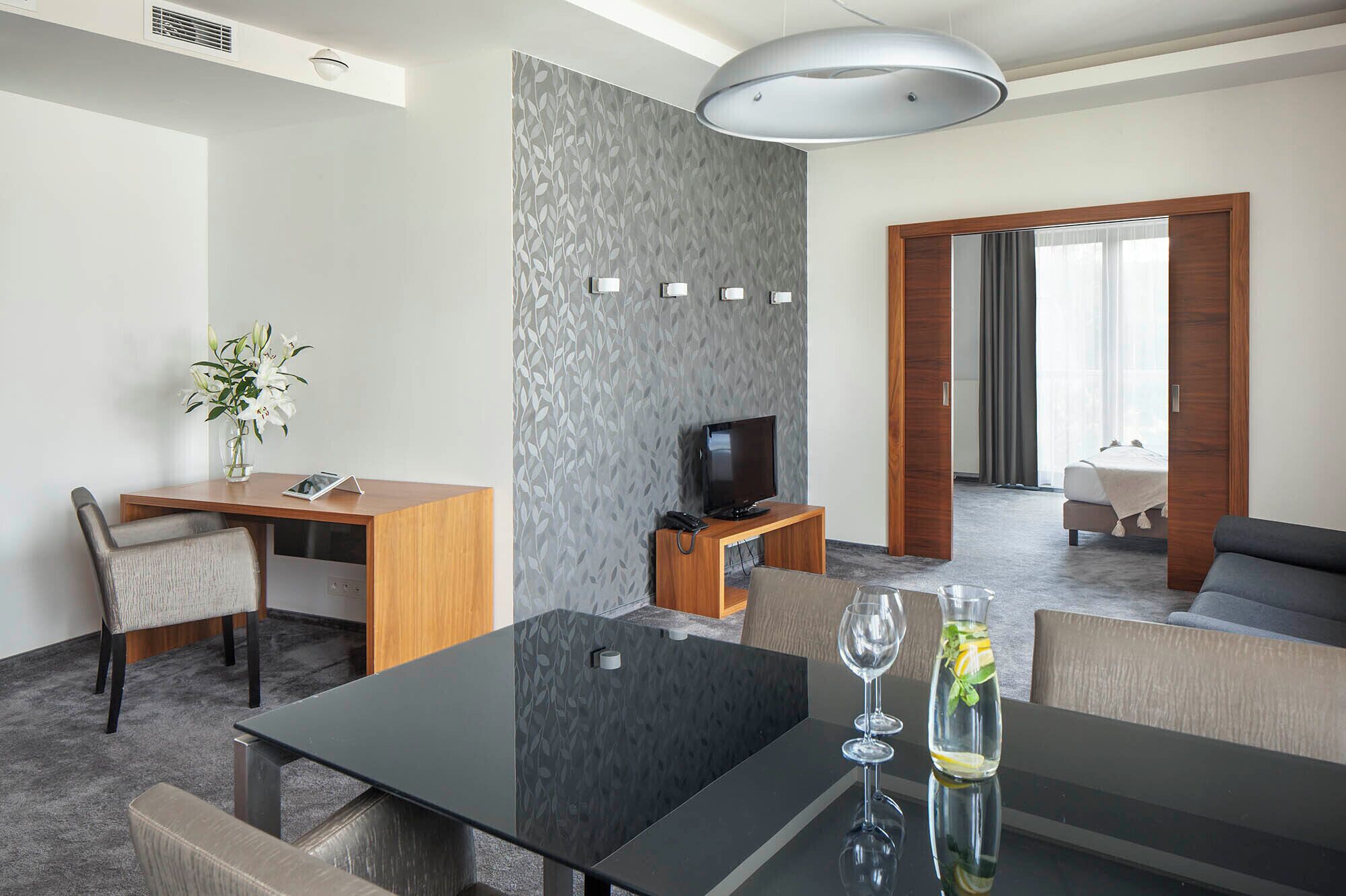 A modern hotel room with a dining area, a TV, a sofa, and a large window.