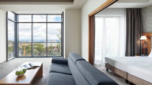 A hotel room with a large window, a sofa, a bed, a bedside table with a lamp, and a view of the cityscape. - Sea Premium Aparthotel (Gdynia)