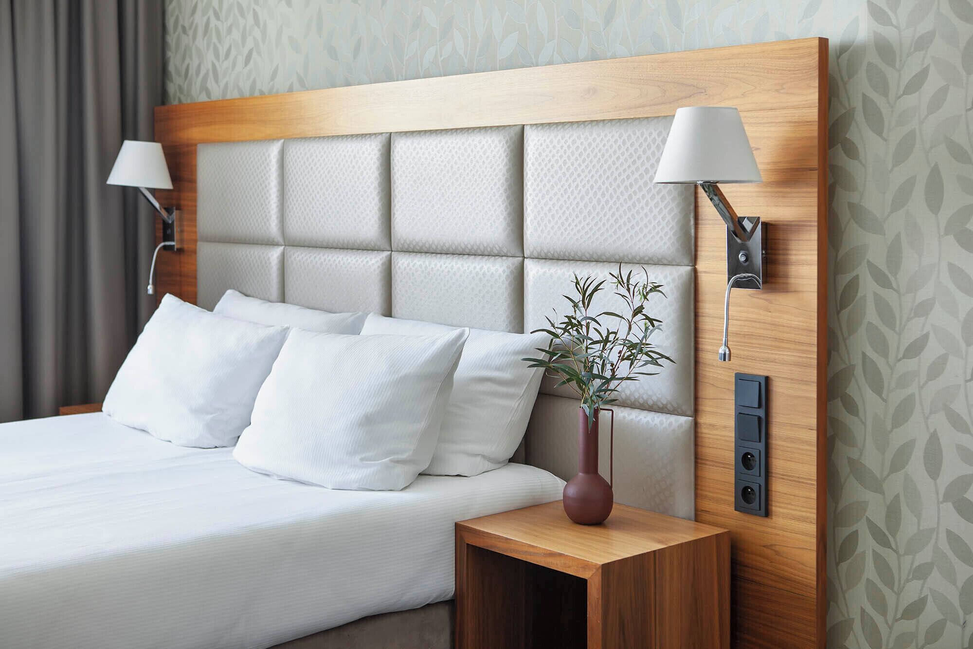 A hotel room with a large bed, two bedside lamps, a wooden nightstand, and a vase with a plant.