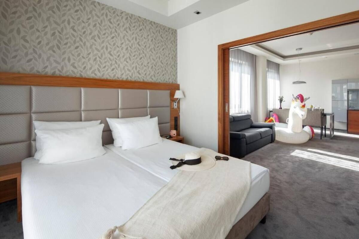 Een hotelkamer met een groot bed, een houten hoofdbord, een nachtkastje, een lamp, een bank en een eetgedeelte zichtbaar door een geopende deur.