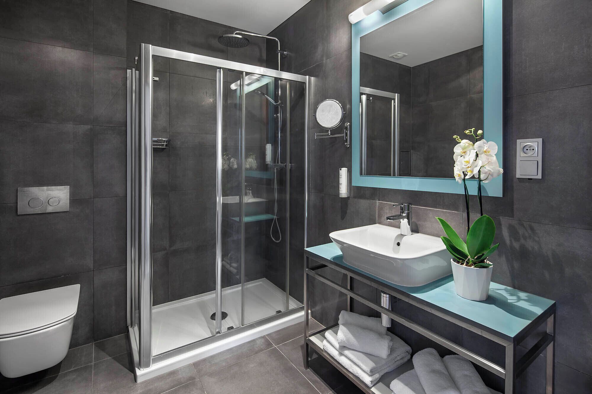 Un baño moderno con ducha de vidrio, un lavamanos blanco, un espejo con marco azul aguamarina y una orquídea en maceta.