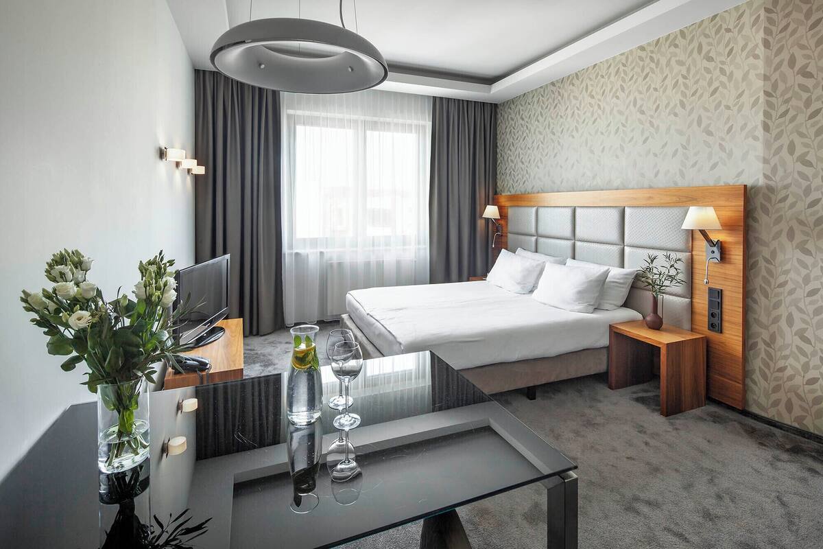 Een hotelkamer met een bed, nachtkastje, lamp, televisie en een glazen tafel met een vaas en glazen.