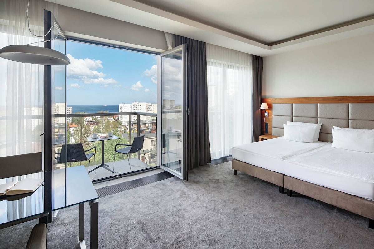 Hotelkamer met een bed, een bureau met stoelen, en een glazen deur die leidt naar een balkon met uitzicht over de stad.