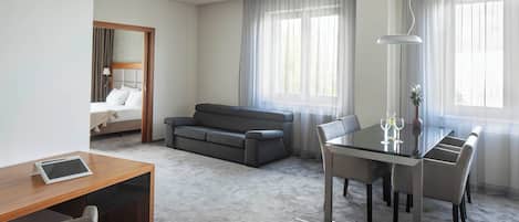 Una habitación de hotel moderna con una cama, un sofá, una mesa de comedor con sillas y un ventanal con cortinas translúcidas.