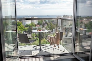 Ein Balkon mit zwei Stühlen, einem kleinen Tisch mit Tasse und Gebäck, und Blick auf Stadt und Meer.