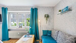 Sala de estar moderna com uma mesa de madeira, cortinas verde-água, um sofá com almofadas estampadas, um radiador e uma janela com vista para o verde.