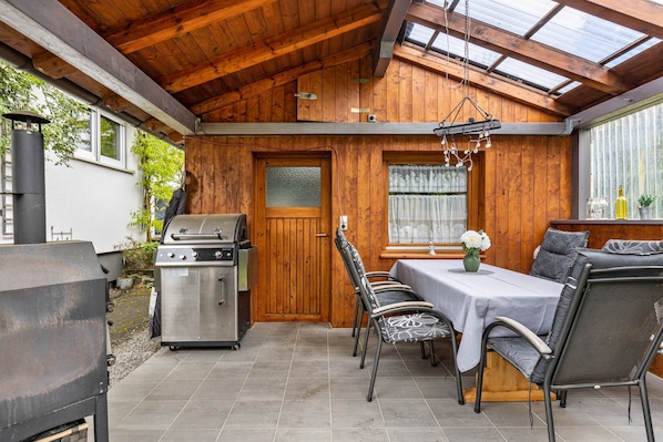 A covered patio with a grill, dining table, chairs, and a view through a window.