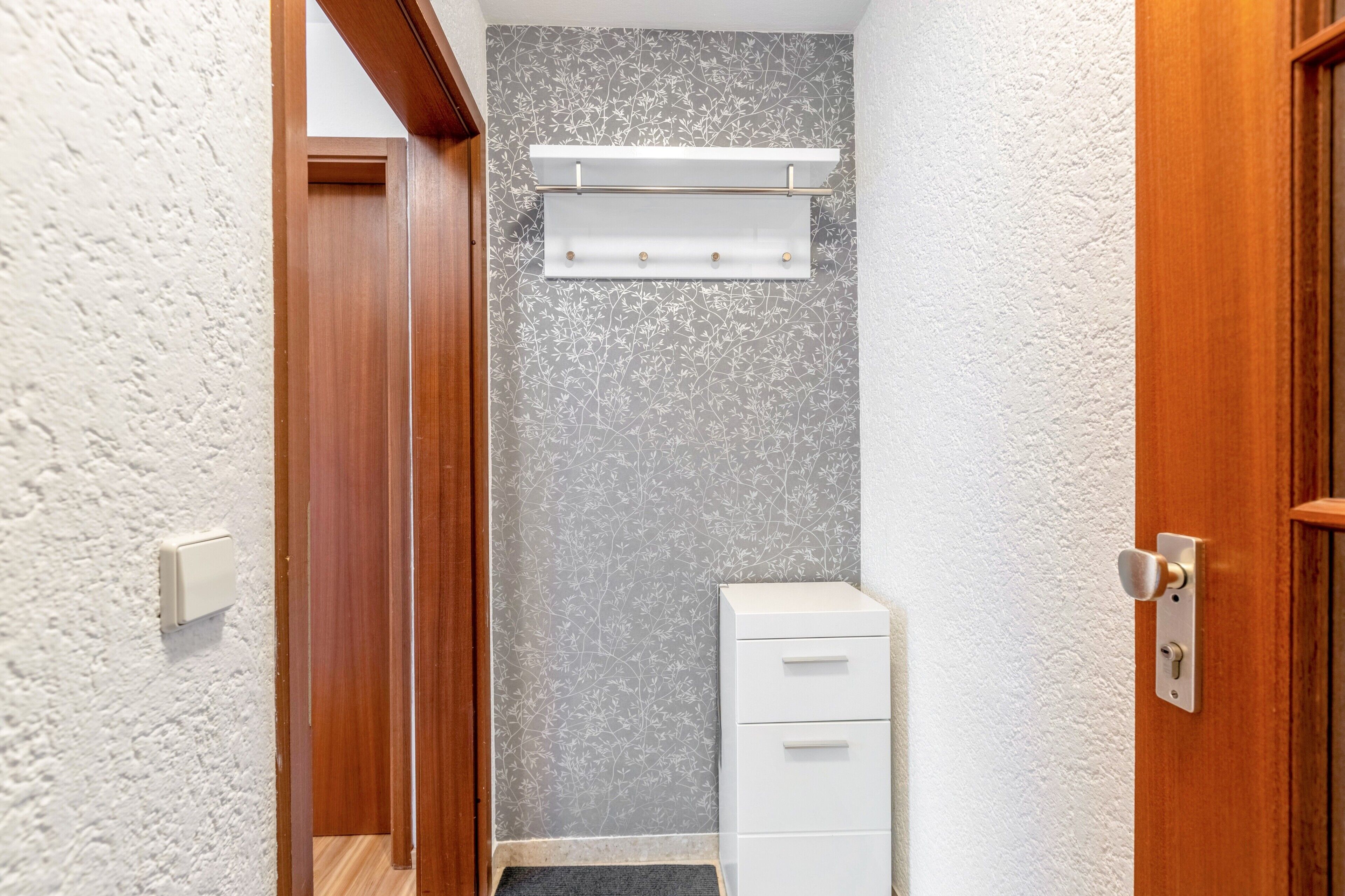 Un couloir avec une porte en bois, un mur blanc orné d’un motif floral, une armoire blanche et une étagère blanche dotée de trois crochets.