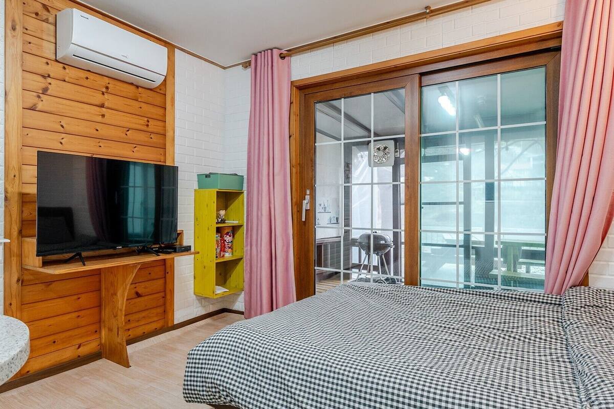 Ein Zimmer mit Fernseher, einem Holzschrank, einem grünen Regal, einem Bett mit karierter Bettwäsche und einer Schiebetür zum Außenbereich.