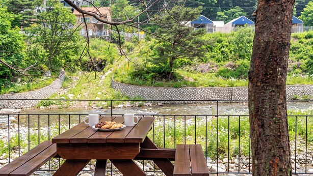 A picnic table with a plate of cookies and two mugs, overlooking a river with a stone wall and greenery. - Gapyeong Funny Meongmeong Dasom (Gapyeong)