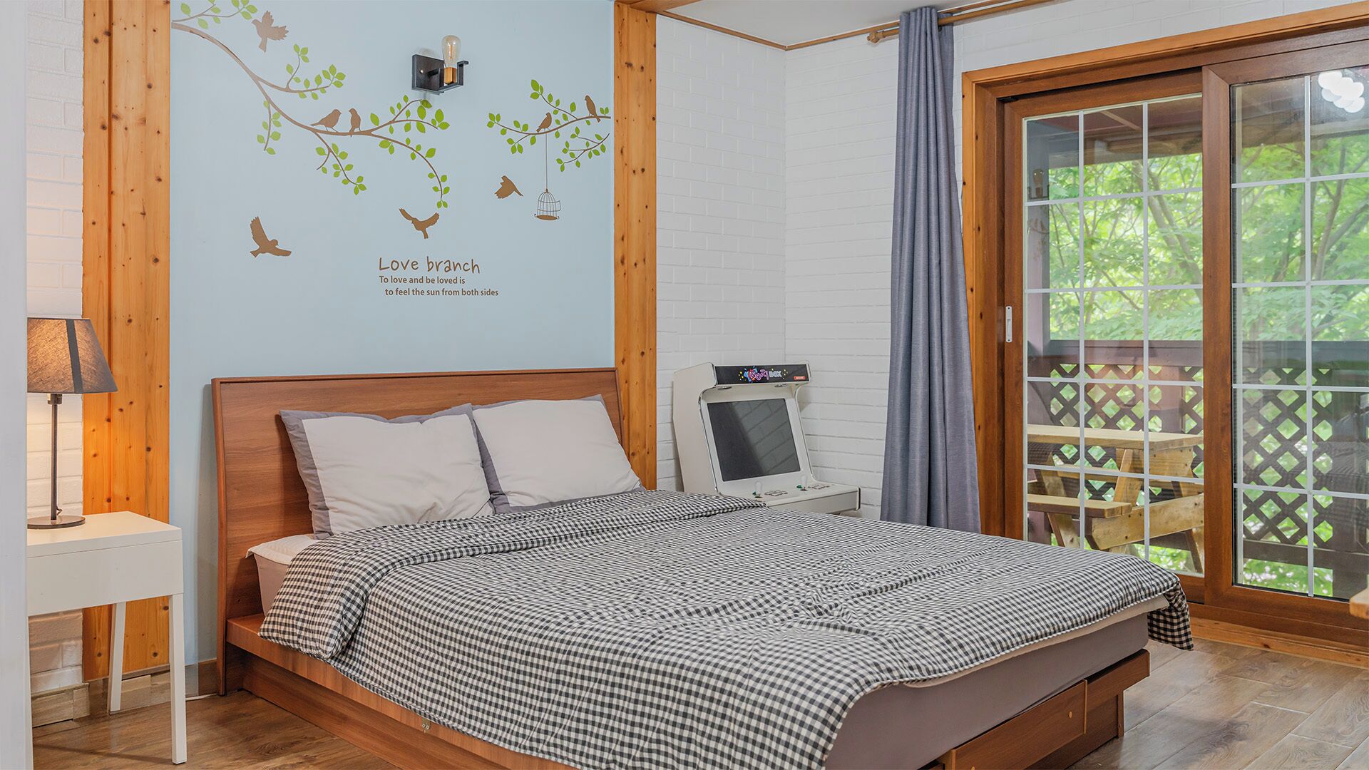 A bedroom with a bed, a wall-mounted lamp, a bedside table, a window with a view, and a wall decal.