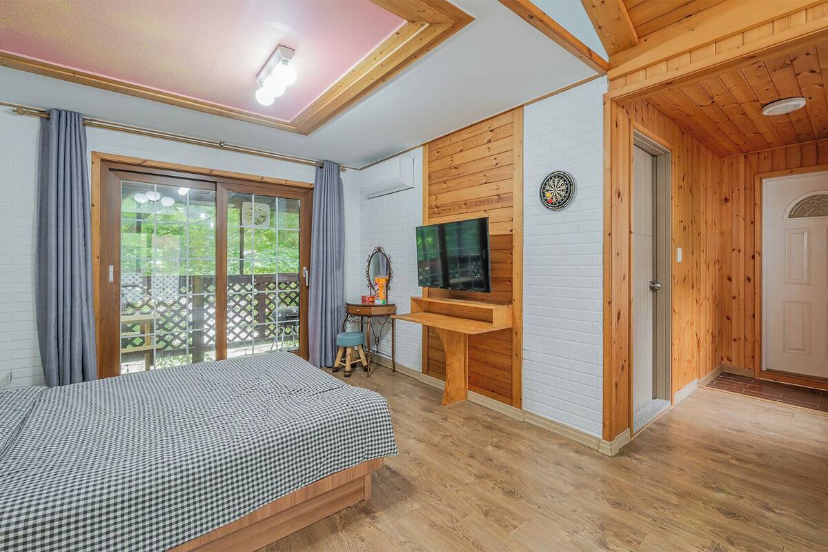 Ein Zimmer mit einem Bett, einem an einer Holzwand montierten Fernseher, einer Tür und einem Balkon mit Blick auf Grünflächen.