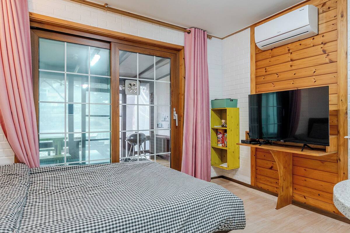 Ein Zimmer mit einem Bett, einem Fernseher, einem Holzschrank, einem Spiegel und einer Glastür zu einem Außenbereich.