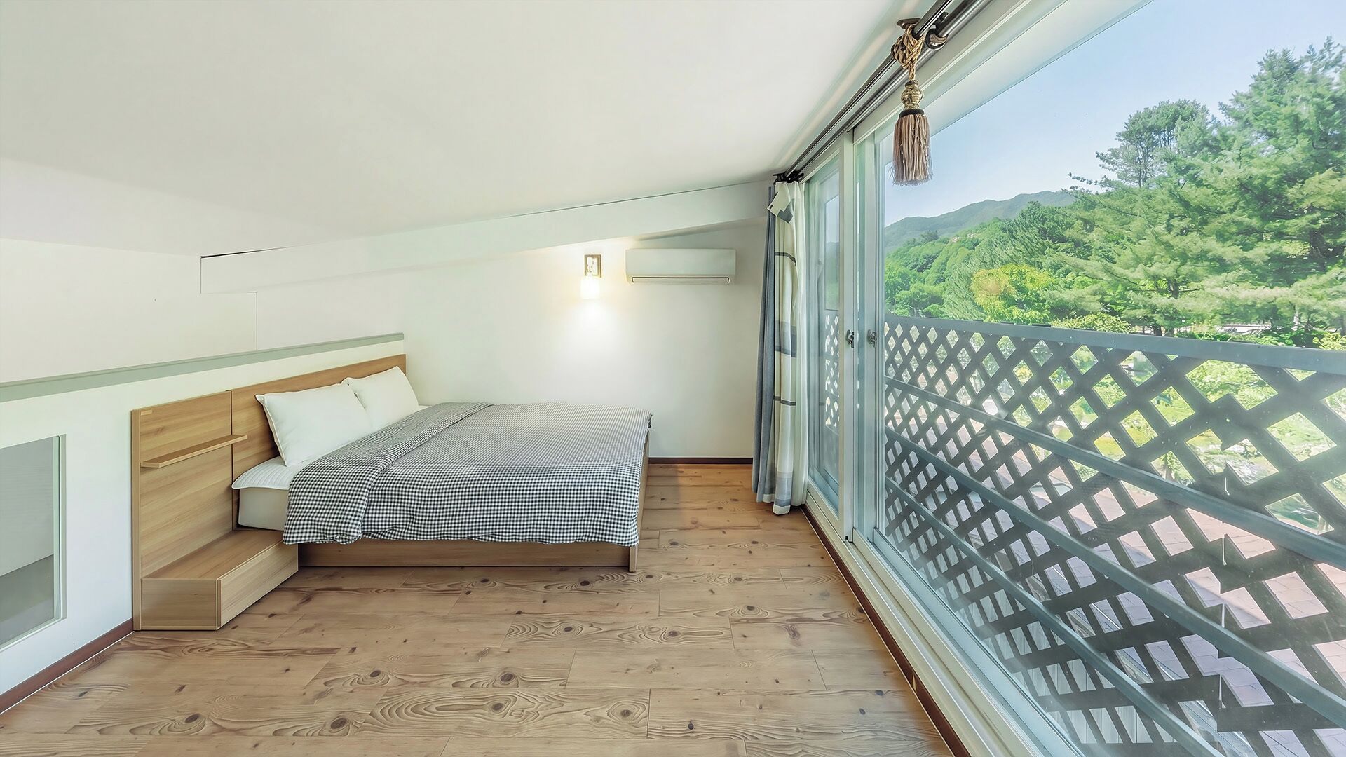 Un dormitorio con cama, suelo de madera, una ventana con vistas a la vegetación y un balcón con barandilla de celosía.