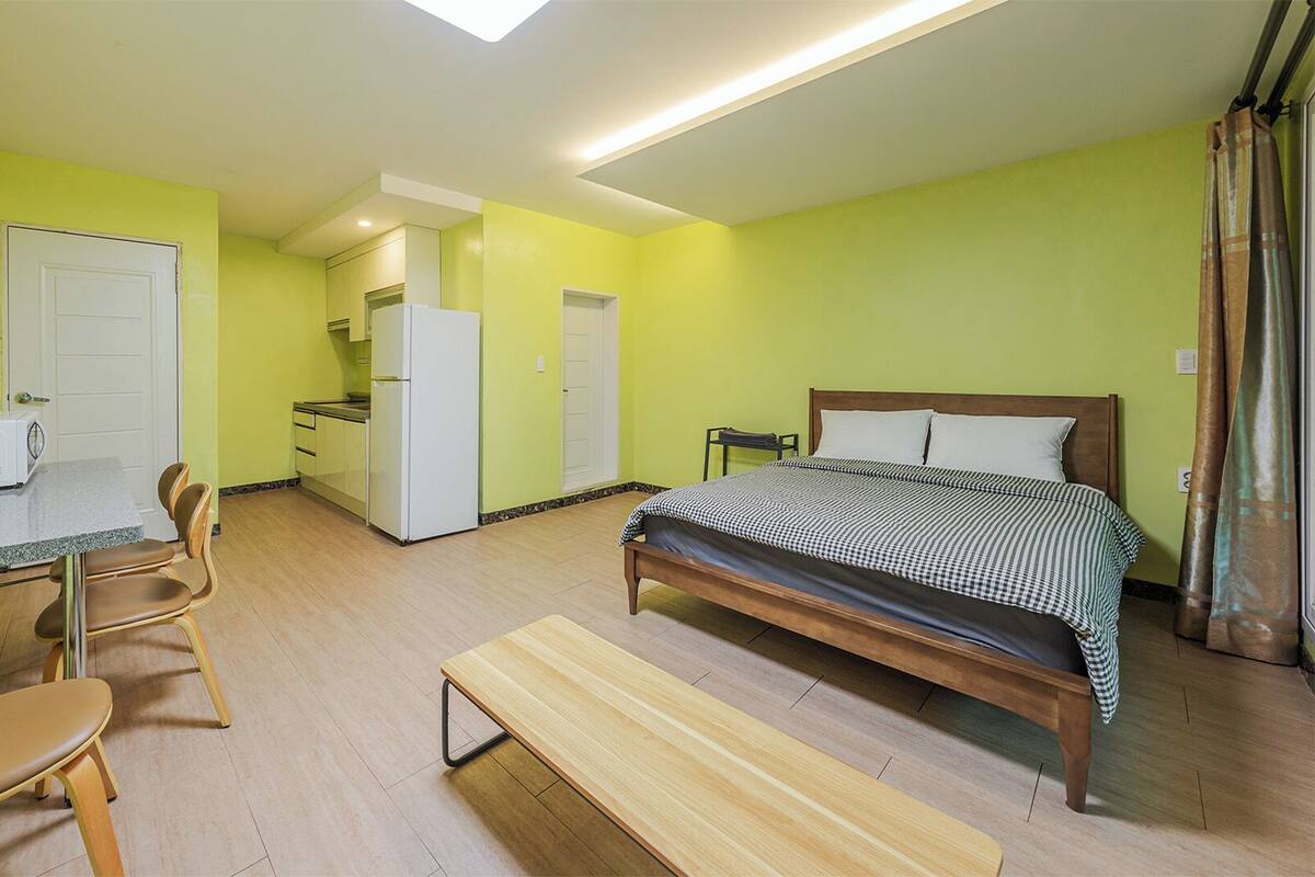 A compact room with a bed, a desk with a chair, a kitchenette, and a bench.
