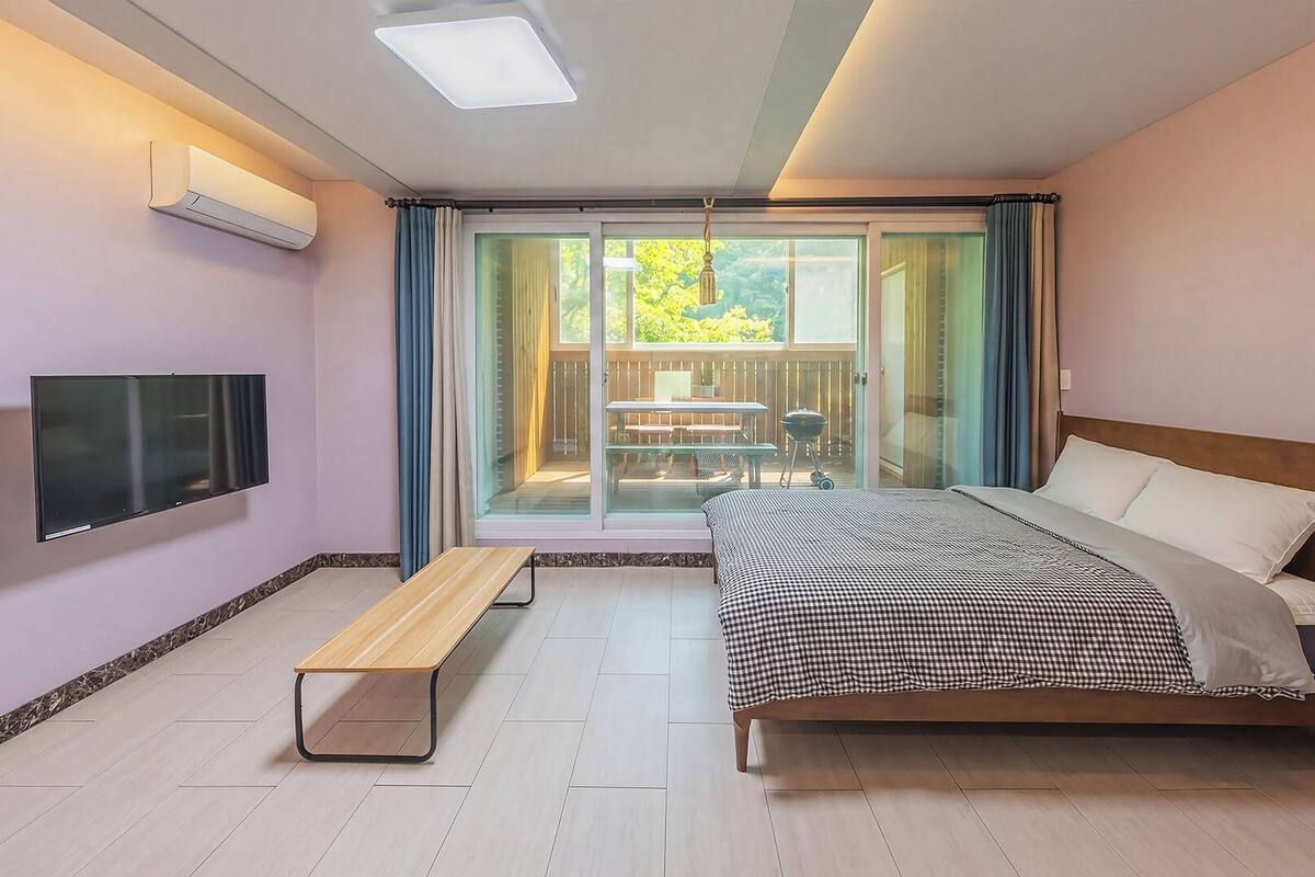 A bedroom with a bed, a bench, a TV, and a sliding glass door leading to an outdoor area.