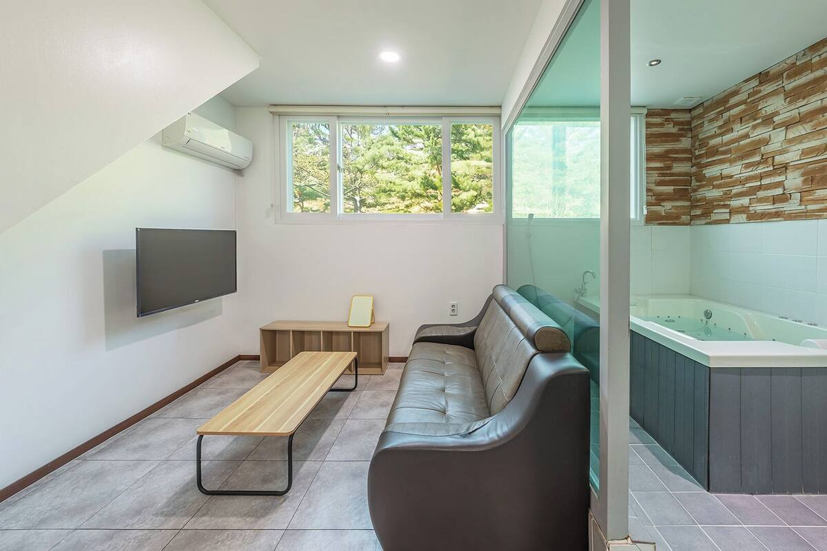 A modern bathroom with a bathtub, a bench, a flat-screen TV, and a window with a view of greenery.