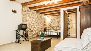 A room with a stone wall, wooden beams, a bed, a small table with a television, and a bathroom visible through an open door. - Vacation home Antonio in the old town of Vrbnik, beautifully furnished (Vrbnik)