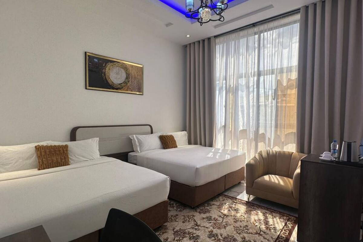 A hotel room with two beds, a sofa, a desk, a chandelier, a large window with curtains, and a framed picture on the wall.