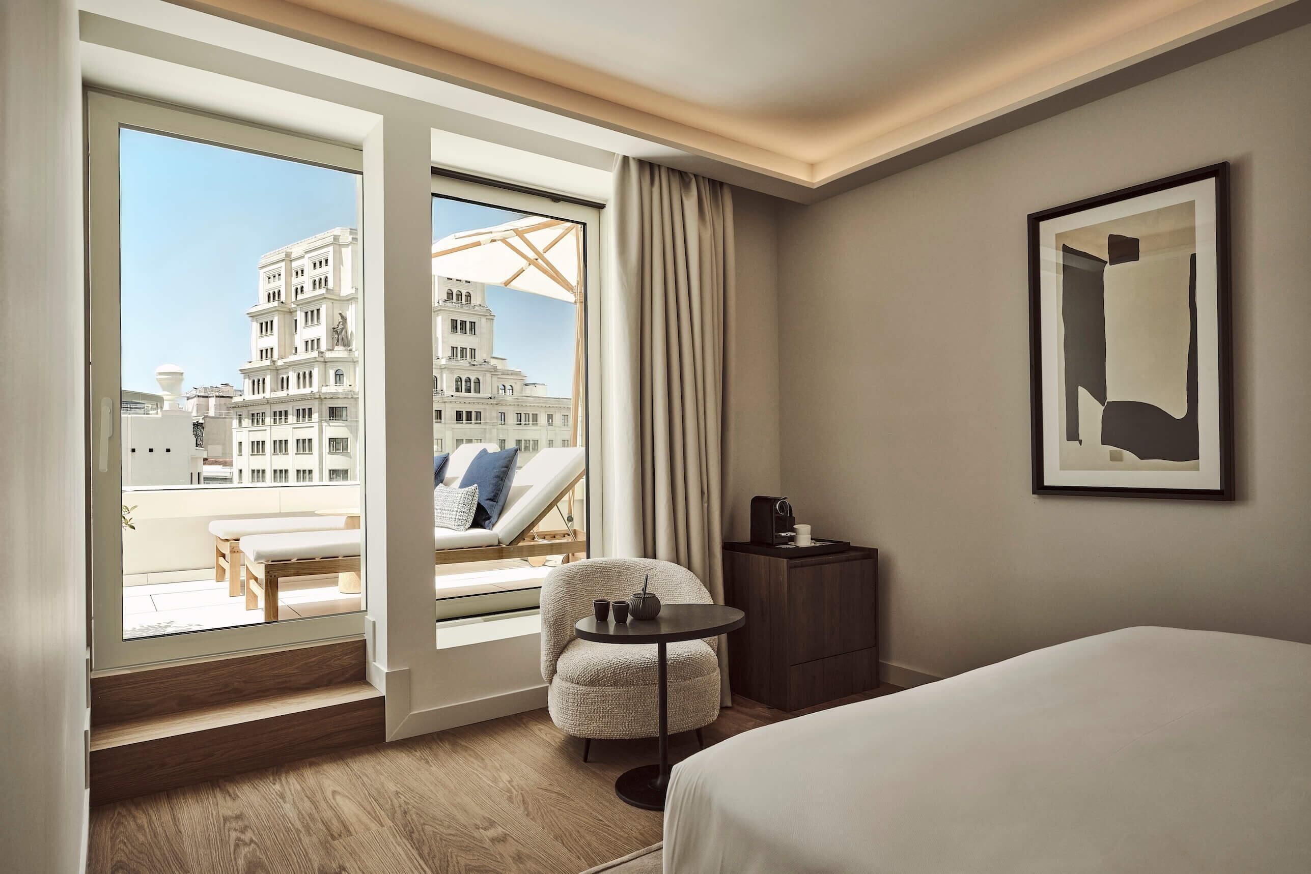 A hotel room with a large window, a bed, a chair, a small table, and a framed picture on the wall.