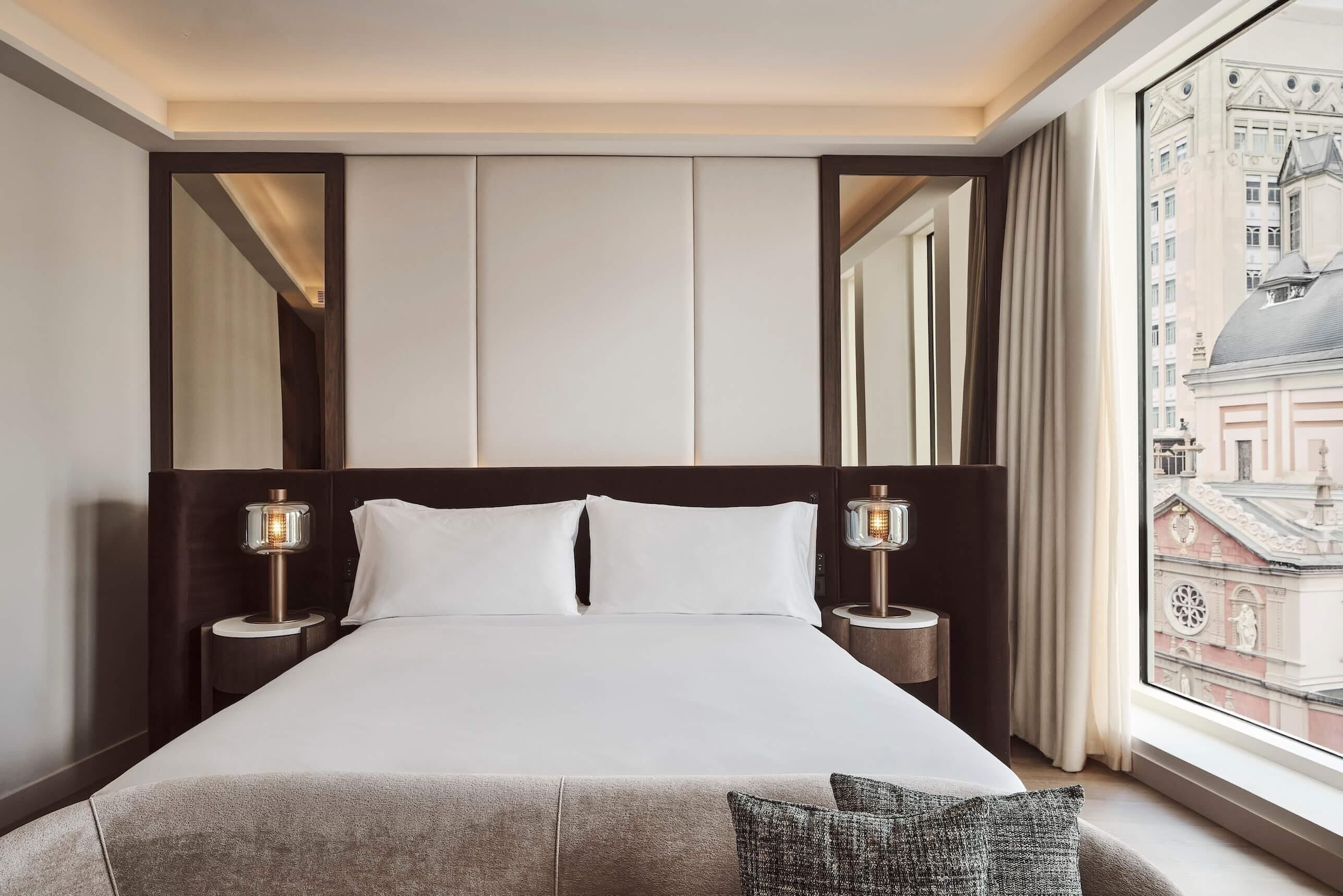 A hotel room with a large bed, two bedside lamps, a window with a city view, and a mirror above the bed.