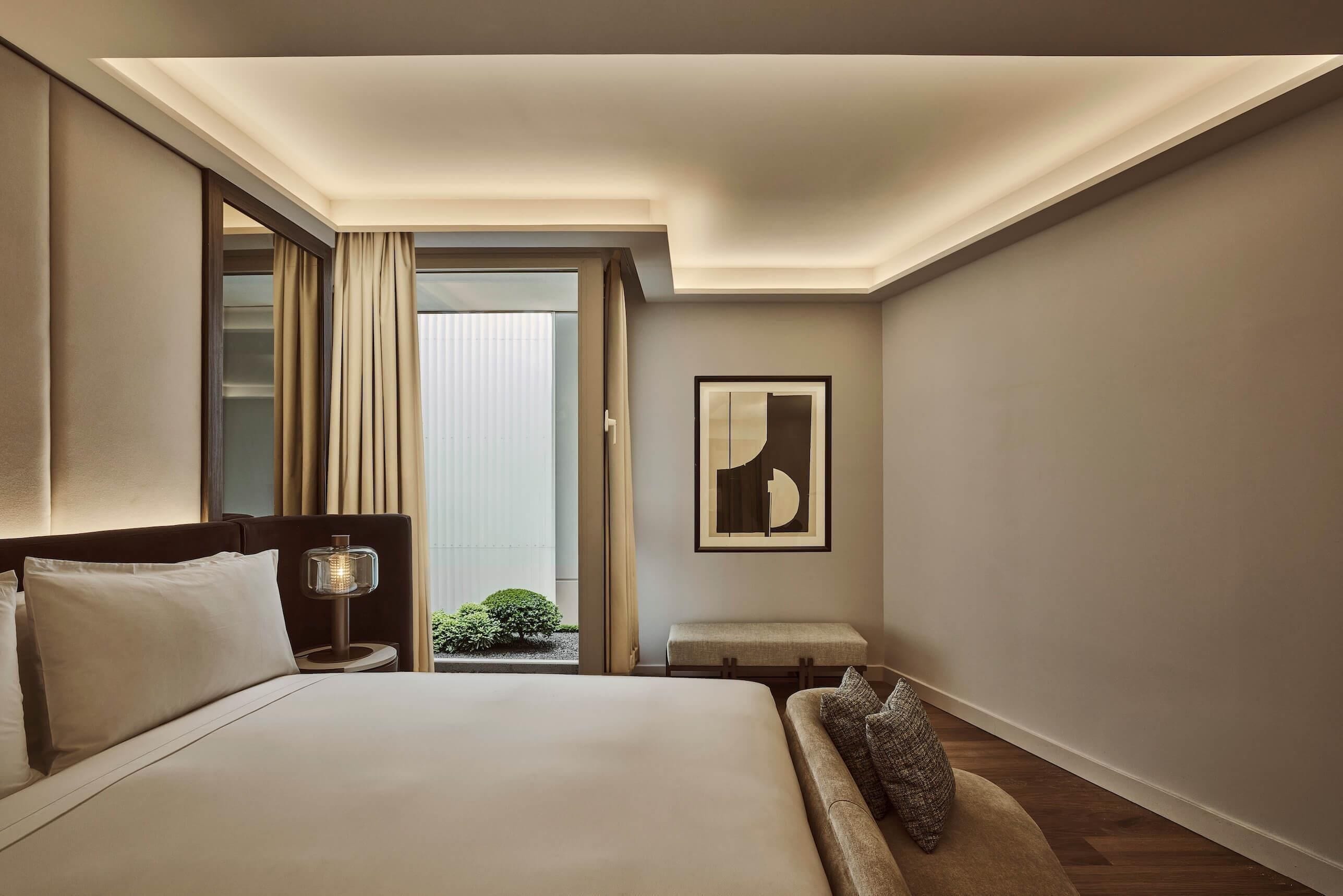 A hotel room with a bed, bedside lamp, bench, and a framed picture on the wall.