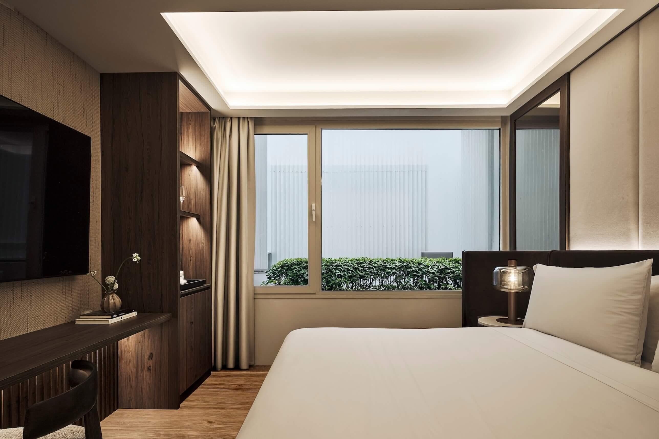 A modern hotel room with a large bed, a desk, a TV, and a window with a view of greenery.