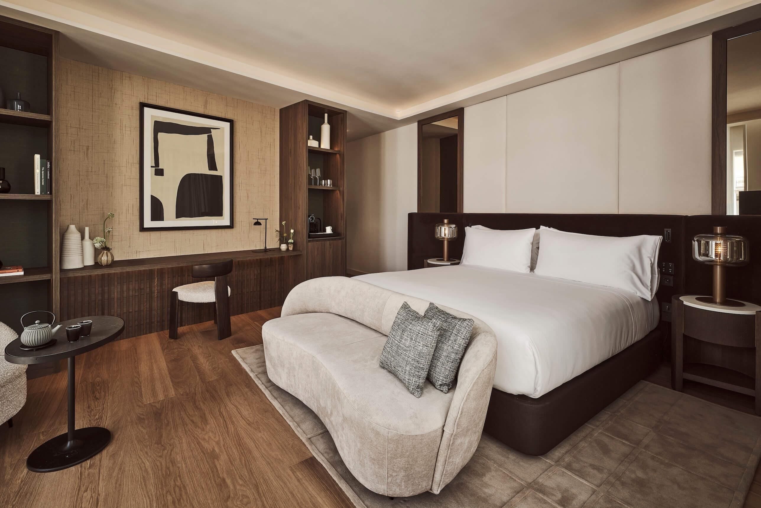 A modern hotel room with a large bed, a sofa, a small table with a kettle, and a bookshelf.