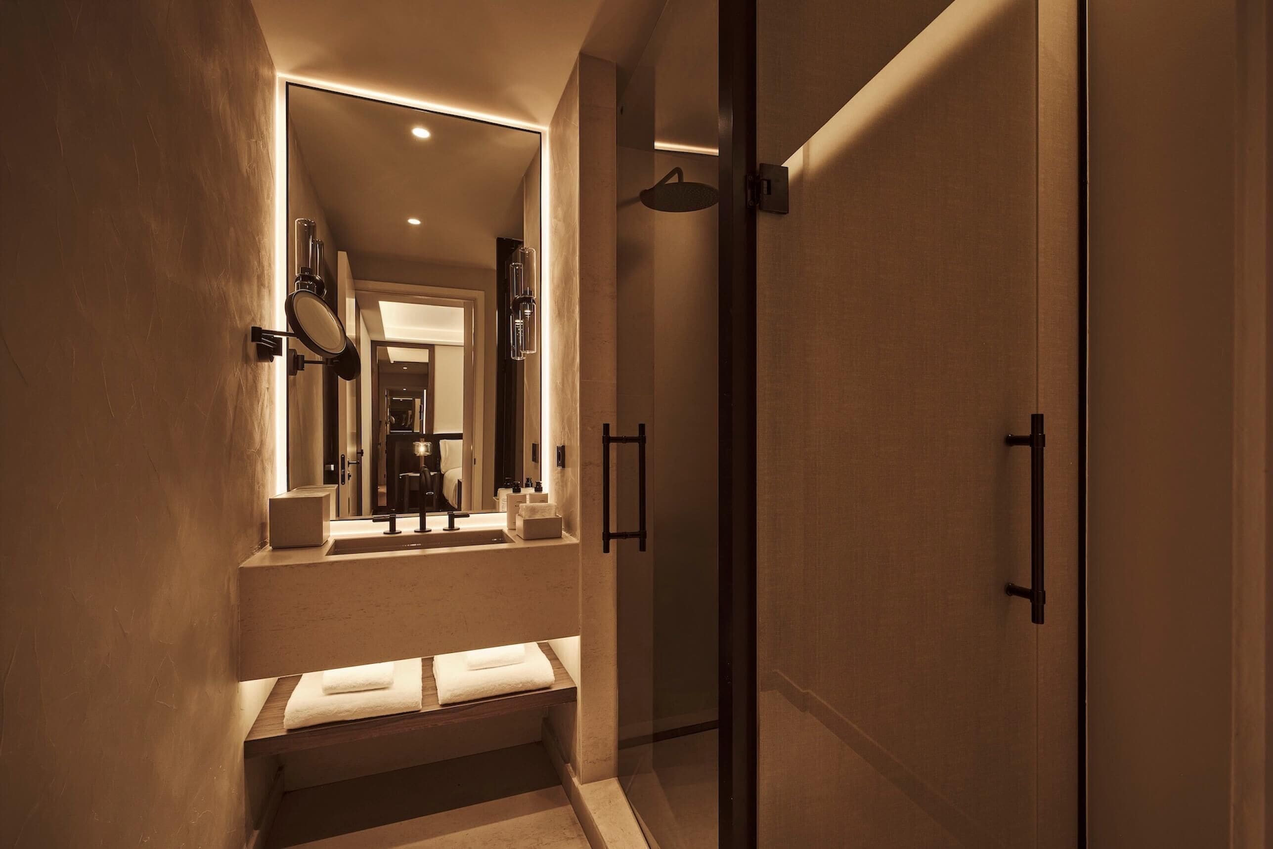 A modern bathroom with a glass shower enclosure, a built-in sink with a mirror, and a bench with towels.
