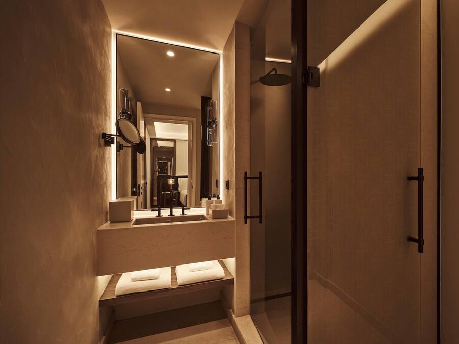 Un baño moderno con cabina de ducha de vidrio, lavabo empotrado con espejo y banco con toallas.