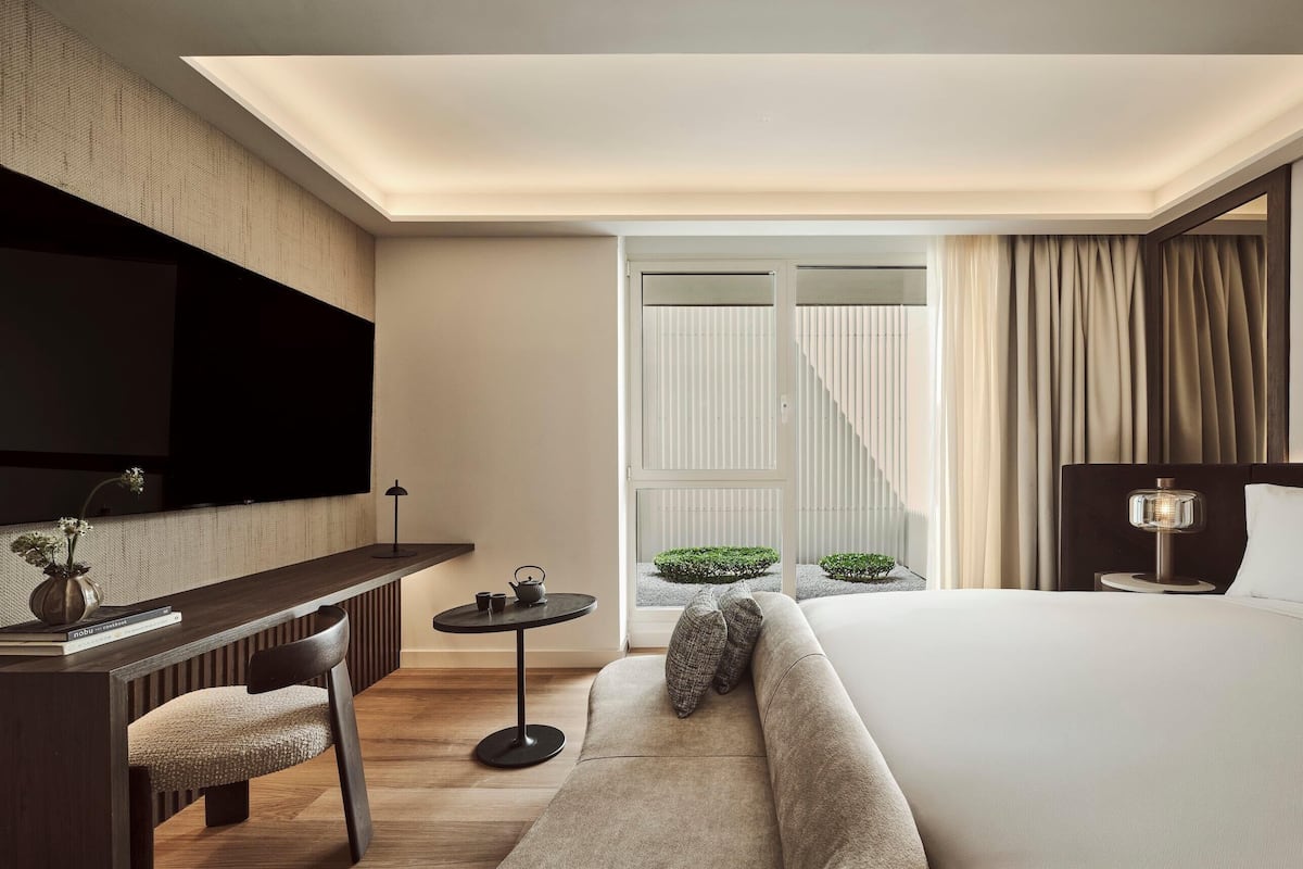 Une chambre d’hôtel moderne dotée d’un grand lit, d’un bureau avec une chaise, d’un téléviseur à écran plat, d’une petite table et offrant une vue sur un jardin grâce à une porte coulissante.