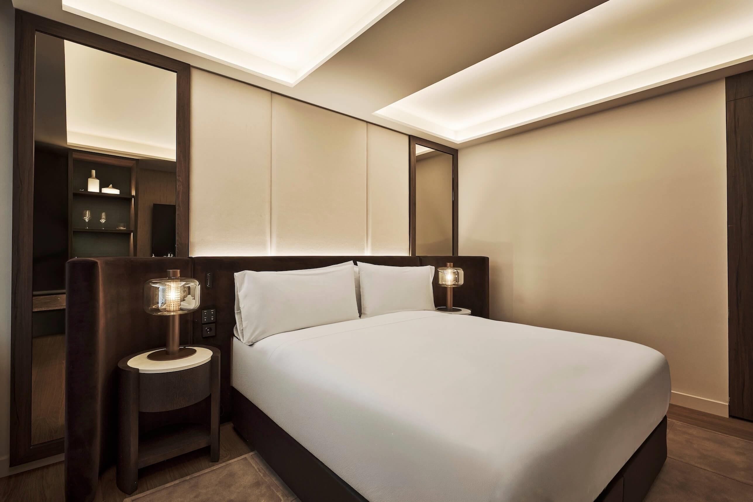 Quarto de hotel moderno com uma cama grande, abajures, uma prateleira embutida e um espelho.