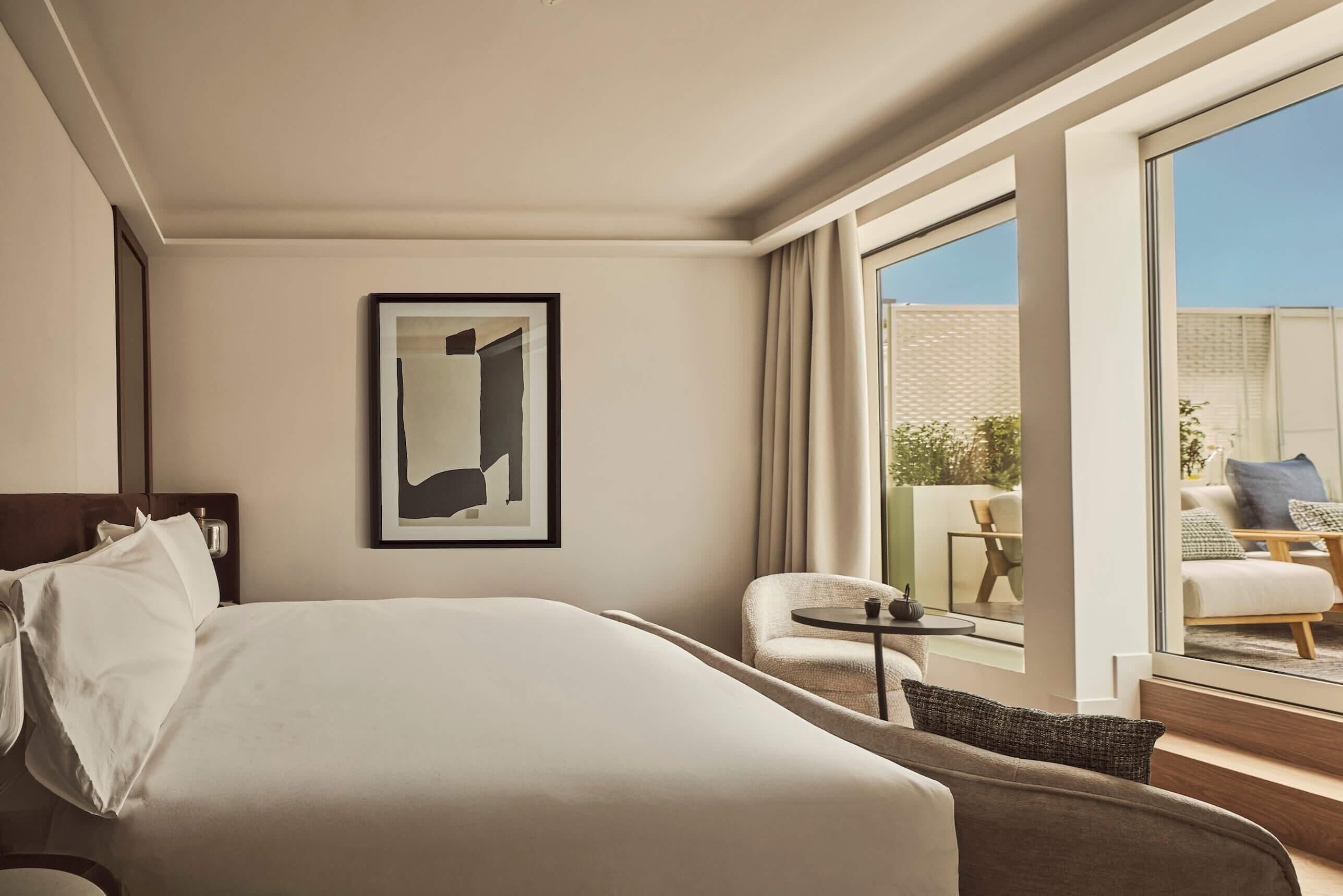 A hotel room with a bed, a small table, a chair, a framed picture, and a balcony with outdoor furniture.