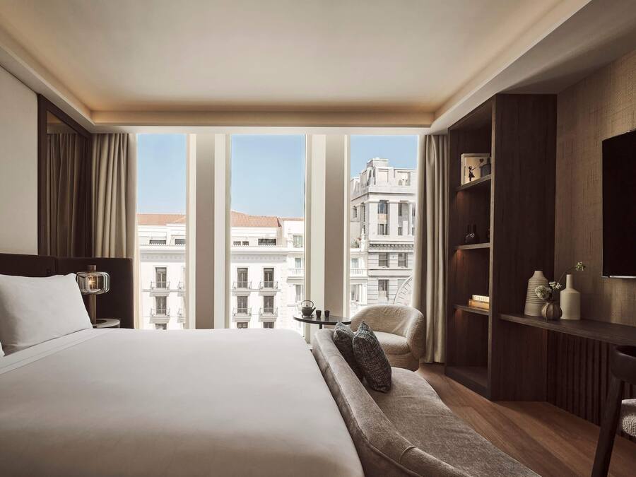 Habitación de hotel con una cama grande, un sofá, un televisor, una mesita de noche con lámpara y vistas a la ciudad.