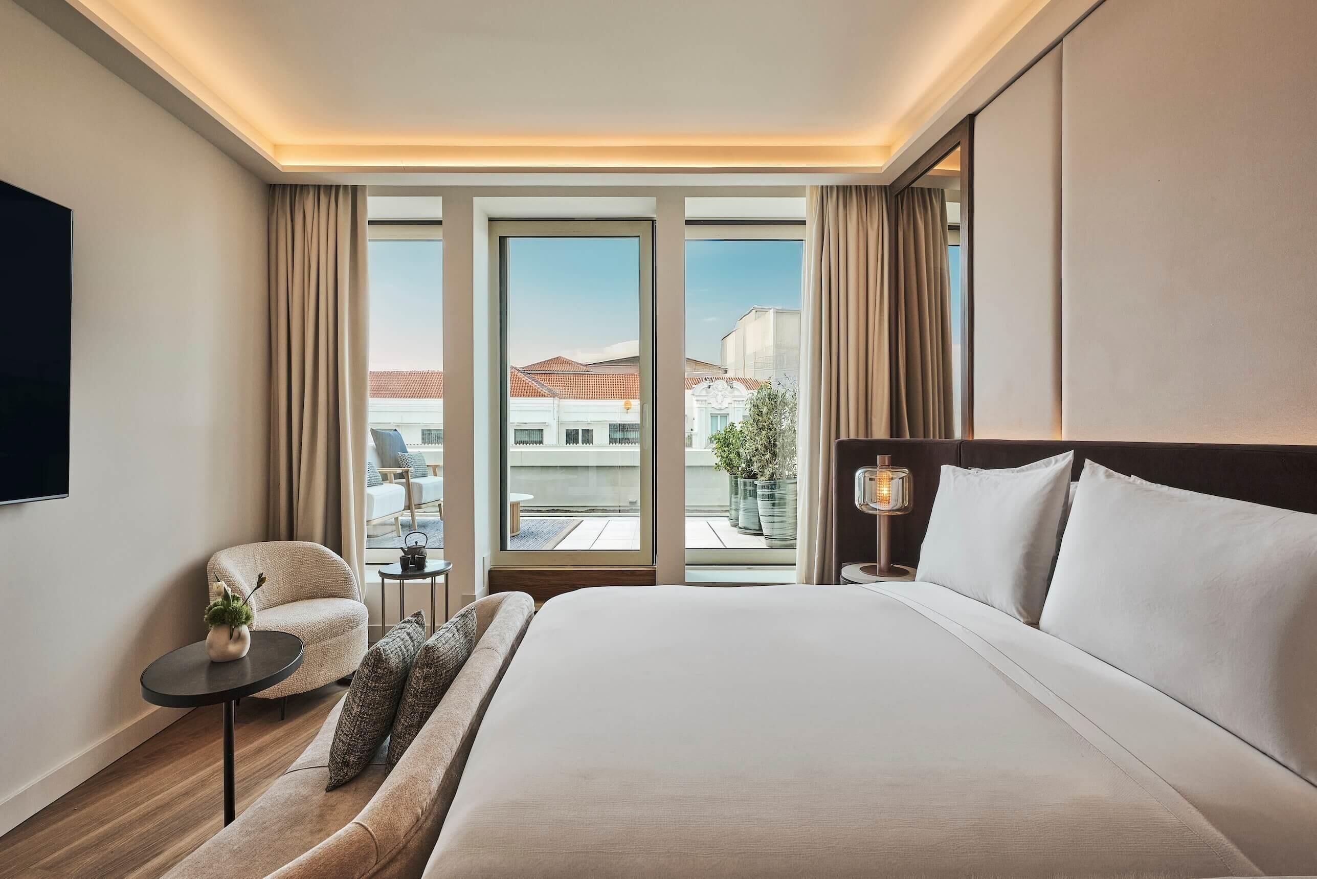 A hotel room with a large bed, a sofa, a small table with a plant, a TV, and a view of buildings through the windows.