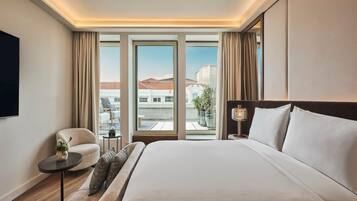 Un cuarto de hotel con una cama grande, un sofá, una mesita con una planta, un televisor y vistas a edificios a través de las ventanas.